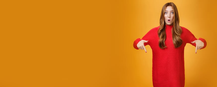 Wow what awesome promotion. Portrait of impressed and surprised cute wondered redhead woman in stylish warm knitted dress folding lips in amazement pointing down and looking at camera photo
