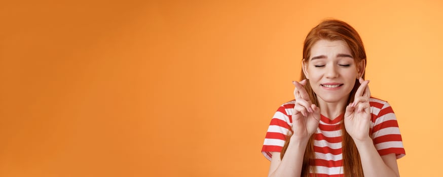 Close-up dreamy cute tender ginger girl make wish, cross fingers good luck closed eyes bite lip eager win lottery, supplicating, praying dream come true, stand orange background believe miracle photo