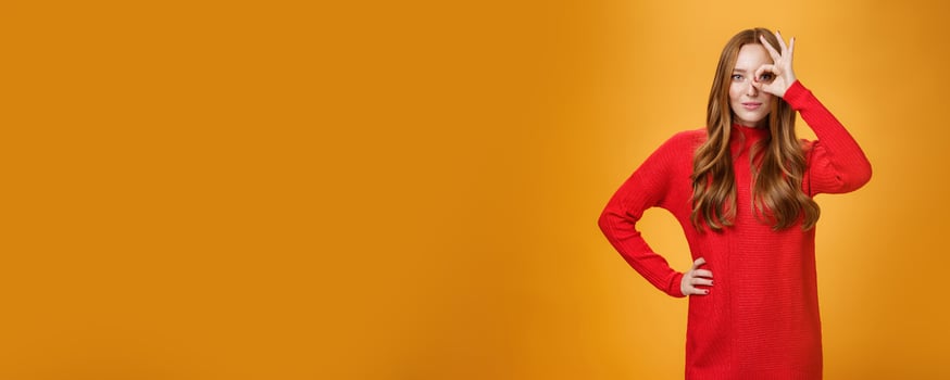 I got you on aim. Self-assured expressive and confident attractive redhead woman in red warm dress showing okay or zero sign on eye and peeking through as smiling satisfied over orange wall photo