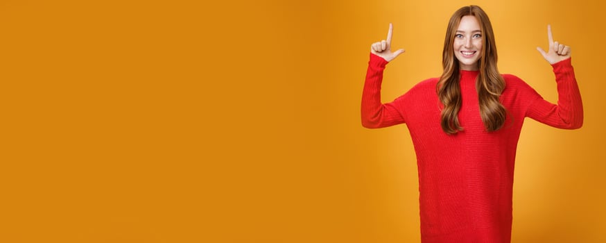 Enthusiastic and excited energized redhead female in red warm sweater pointing up with rised hands and smiling broadly thrilled with awesome promotion, feeling astonished against orange background photo