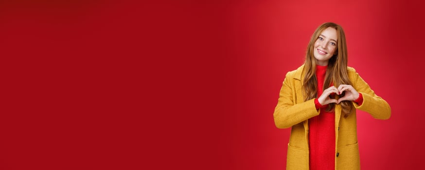 Love, romance and fall concept. Portrait of charming tender and gentle young redhead woman in yellow coat showing heart gesture making confession in sympathy, smiling cute at camera over red wall photo