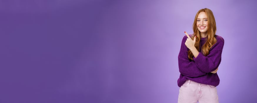 Charming friendly-looking bright redhead woman with freckles and makeup in purple warm sweater pointing at upper left corner and smiling delighted and cute at camera as showing cool place hang out photo