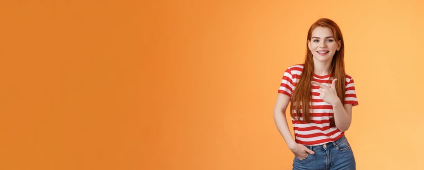 Carefree modern 20s female student chatting friends discuss left copy space, pointing left relaxed casual pose, smiling camera joyfully, introduce advertisement product, orange background photo