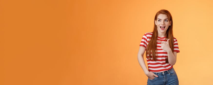 Enthusiastic amazed sassy modern redhead female attend impressive bad awesome design, look amused, observe cool place, pointing left excited, open mouth fascinated intrigued, stand orange background photo