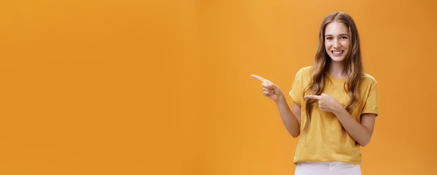 Girl shows cool place to visit. Portrait of charming carefree pretty young lady with cute natural wavy hair pointing left with index fingers and smiling broadly howing perfect place for advertisement photo