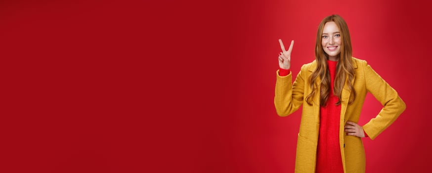 Portrait of happy playful and cute redhead woman with freckles in yellow coat holding hand on waist confident and self-assured showing peace or two sign delighted, beating weather conditions photo