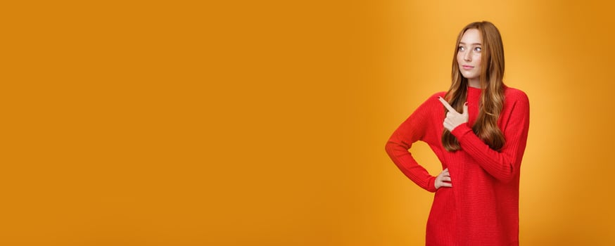 Portrait of glamour and stylish redhead female entrepreneur designing and leading new project pointing at upper left corner as gazing thoughtful and focused on job, managing business over orange wall photo