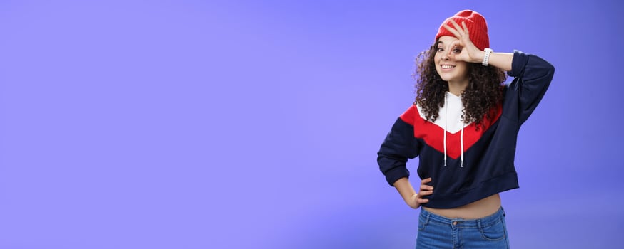 Okay girl agrees. Friendly-looking happy and charming young curly-haired girl in warm beanie showing ok gesture or circle over eye as peeking at camera and smiling broadly with joy over blue wall photo