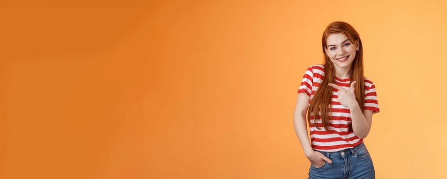 Tender cute feminine redhead lovely girl, tilt head cheerfully smiling, pointing left, showing way, give advice what purchase, advertising good product, introduce shop, orange background photo