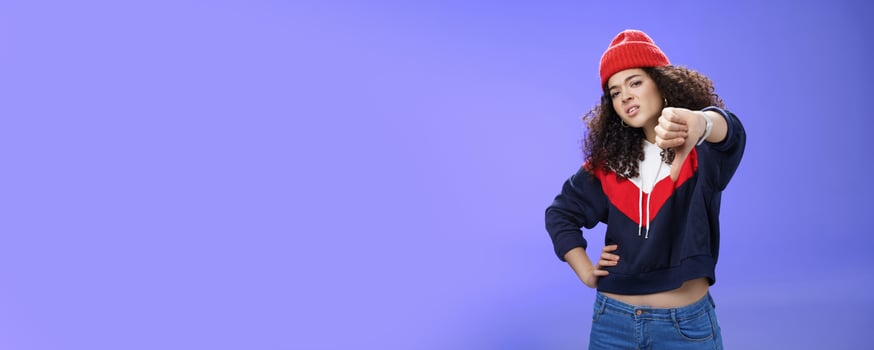 Nah dislike. Portrait of annoyed and unimpressed arrogant cool girl with curly hair tilting head with distain and scorn showing thumbs down in disappointment grimacing, posing over blue wall photo
