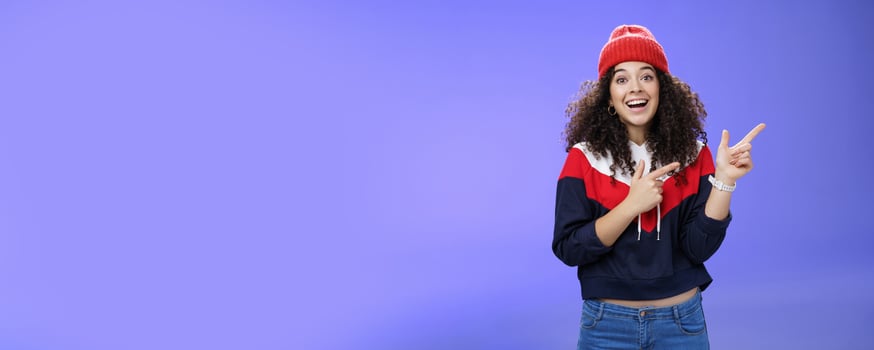 Enthusiastic friendly-looking attractive young 20s woman with curly hair in warm beanie and sweatshirt smiling open mouth amazed and delighted as pointing at upper right corner over blue background photo