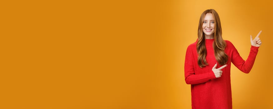 Friendly-looking joyfuly and energized redhead female in red sweater pointing at upper right corner promoting advertisement with broad delighted and tender smile posing against orange background photo