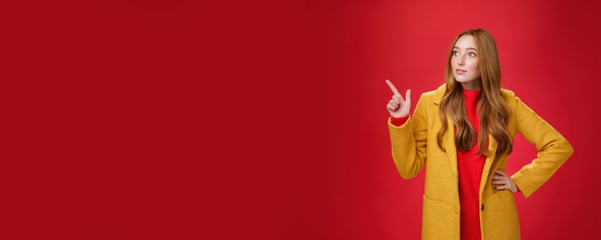Wondered curious good-looking redhead woman in coat stopping on way looking and pointing at upper left corner curious and interested, observing surprising promotion over red background photo