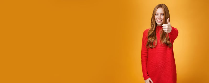 Nice job good. Portrait of delighted and energized confident ginger woman in red sweater pulling thumb up at camera and smiling, liking idea, approving and accepting it over orange background photo