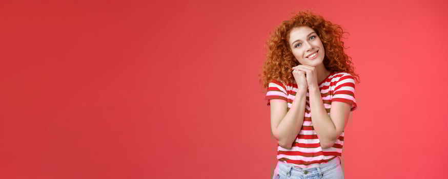 Tender feminine caring redhead curly-haired female romantic gaze camera tilt head clench palms near head silly lovely look smiling delighted cheerful dreamy sighing look with sympathy photo