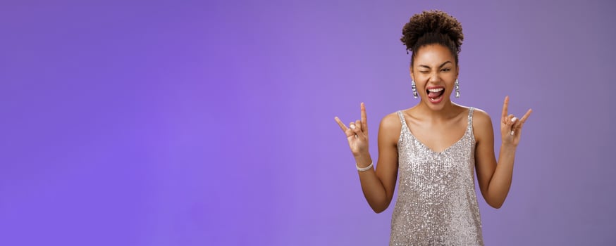 Excited good-looking african-american woman in glittering silver shiny dress having fun amusing university party prom night show tongue winking joyfully rock-n-roll heavy-metal gesture, blue wall photo