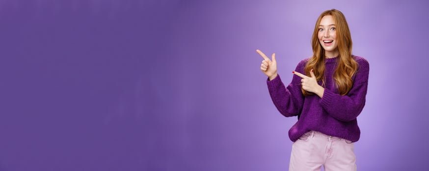 Come in. Portrait of friendly-looking cheerful pretty ginger girl in purple sweater inviting us visit place pointing left with finger pistols and smiling broadly with happy expression over violet wall photo