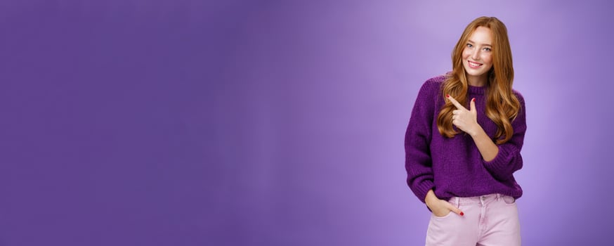 Glamour and stylish feminine woman with long natural red hair in purple sweater holding hand in pocket as pointing at upper left corner showing place she does hairstyle or makeup over violet wall photo