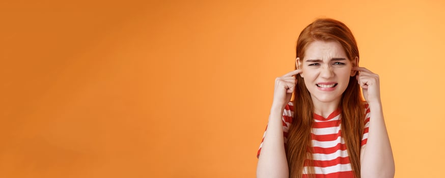Turn off sound. Annoyed disturbed redhead female cannot study hear bothering loud noise, plug fingers ear holes grimace, clench teeth displeased, sibling awful play violin, orange background photo