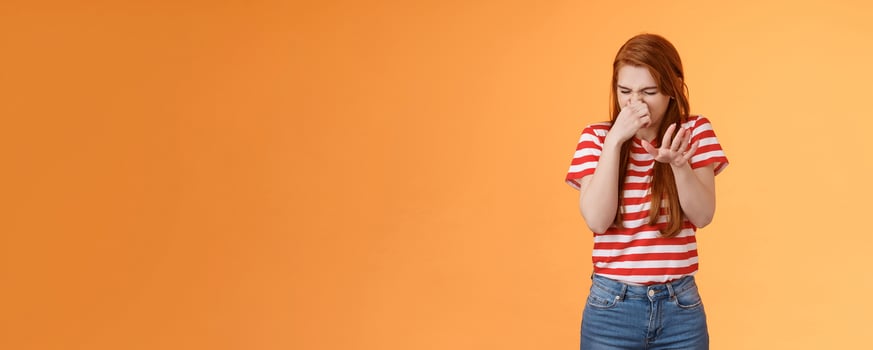 Gosh it stinks. Reluctant disgusted redhead woman raise hand block dislike gesture shut nose, cringe from aversion dislike, puke from reek bad smell, stand orange background disappointed photo