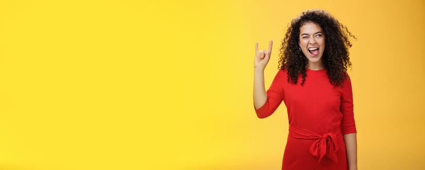 Rebellious meloman leaving in tender girl. Thrilled and carefree curly-haired woman in red dress sticking out tongue and looking right with smile as showing rock-n-roll gesture, enjoying cool music photo