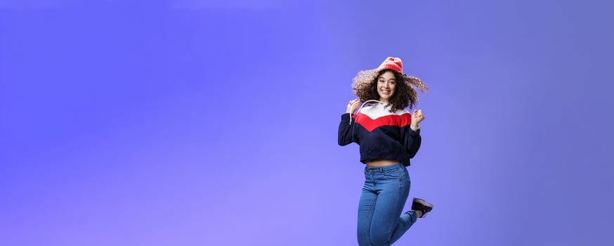 Delighted carefree and happy young woman with curly hair in warm beanie jumping joyfully having fun raising hands and smiling broadly posing over blue background in outdoor clothes photo