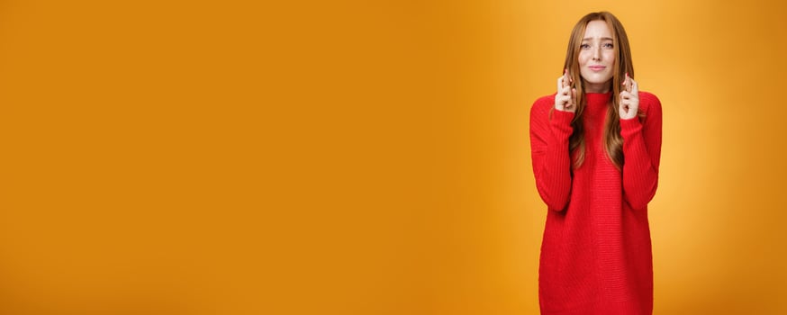 Hopeful worried redhead girl in red sweater frowning pursing lips cross fingers for good luck, supplicating and praying for dream come true, standing excited against orange background photo