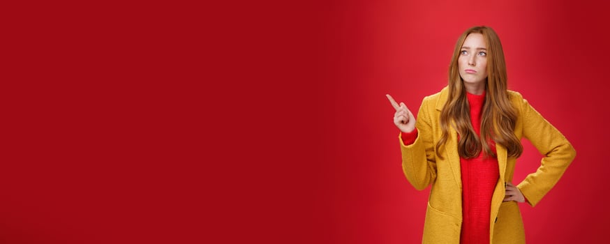 Sad and gloomy cute ginger girl in 20s wearing trendy yellow coat holding hand on waist, pouting, looking and pointing at upper left corner with regret and sorrow over red background photo