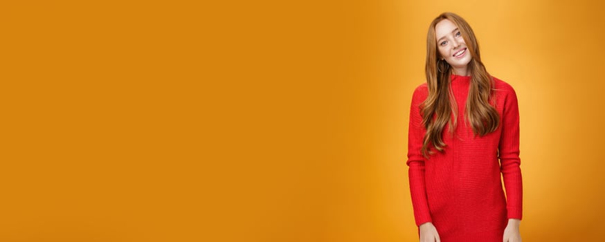 Attrative and stylish elegant busiensswoman with ginger hair in red knitted dress tilting head and smiling flirty and happily posing over orange background as listening with interest to conversation photo