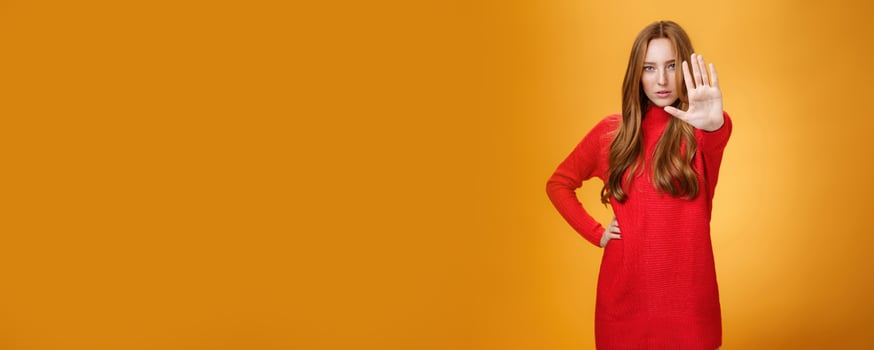 Confident sensual and attractive redhead female with self-assured gaze extending palm towards camera in stop and prohibition gesture demanding halt, stand still over orange background photo