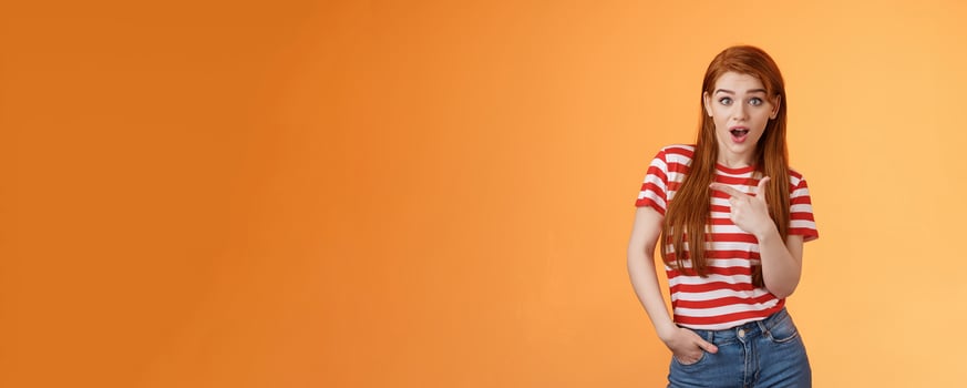 Outgoing good-looking redhead modern girl wonder making amazing purchase, look camera surprised, questioned pointing left, interested about new cool promo, stand orange background photo