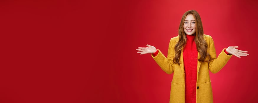 Redhead girl apologizes for being late smiling silly and guilty with sorry look shrugging as raising hands sideways wearing stylish yellow coat over dress posing against red background photo