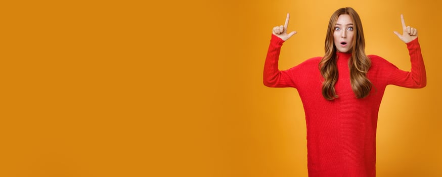 Portrait of attractive and stylish redhead 25s female in knitted red dress gasping from amazement open mouth questioned and surprised as pointing up at astonishing promotion over orange wall photo