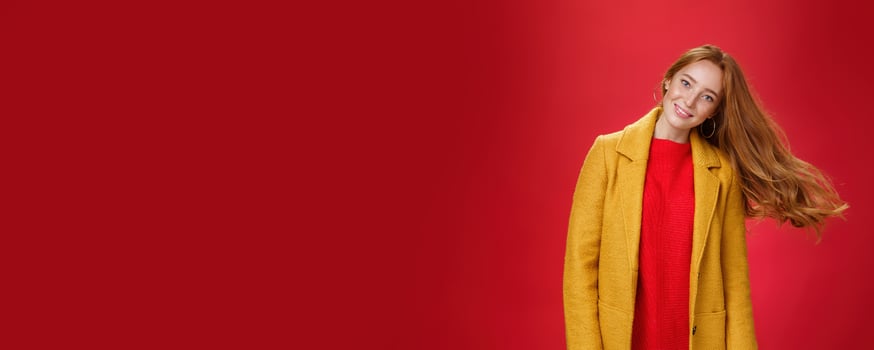 Charming redhead female with freckles waving hair smiling broadly tilting head as haircut flying in air posing delighted and carefree over red background in warm yellow coat and knitted dress photo