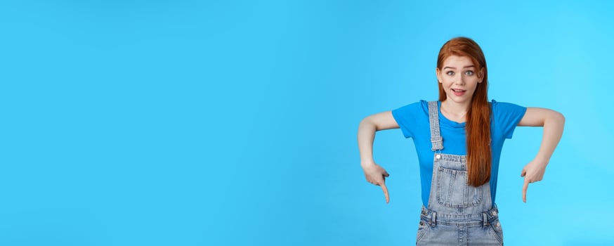 Curious tender lovely ginger caucasian girl, pointing down questioned, check out interesting promo, point bottom down copy space, look camera intrigued, suggest good product online shop link photo
