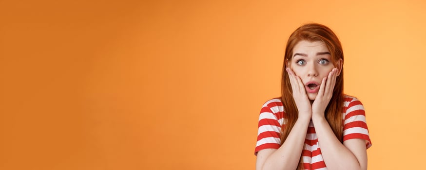 Close-up shocked speechless redhead woman, drop jaw gasping, touch cheeks embarrassed shook, look pity shame, stare camera astonished, surprised hear disturbing news, orange background photo