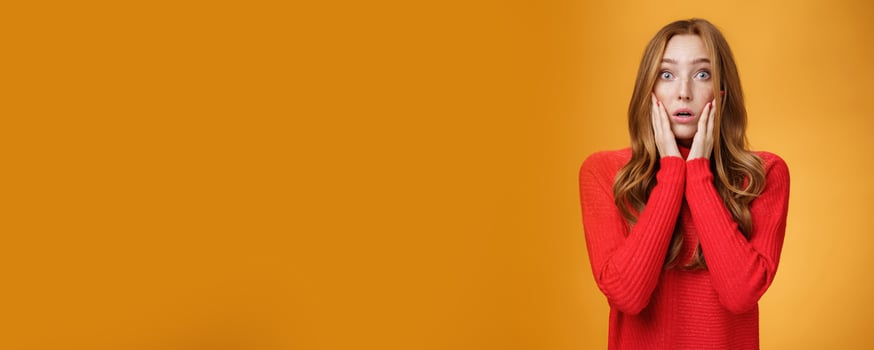 Worried shocked redhead female reacting to stunning troublesome news holding palms on cheeks open mouth and raise eyebrows astonished, posing concerned against orange background photo
