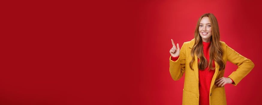 Charming young female in yellow coat holding hand on waist and pointing at upper left corner, smiling broadly as showing or promoting interesting proposal over red background, in good mood photo
