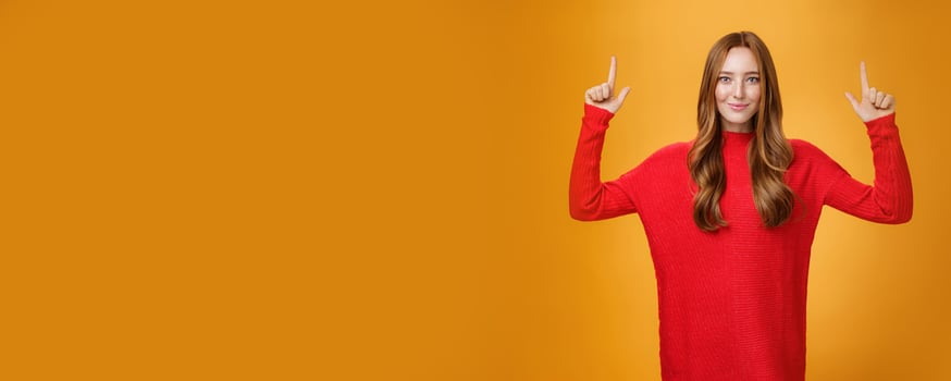 Look up you not regret it. Portrait of confident and stylish young redhead woman in 20s wearing knitted red dress pointing upwards and smiling assertive with self-assured grin over orange background photo