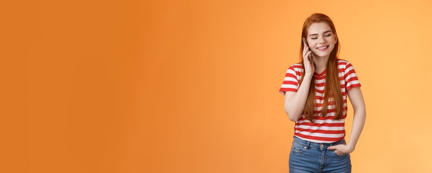 Friendly talkative good-looking redhead female chatting lively discuss ordinary things hold smartphone near ear, close eyes smiling joyfuly, communicating, listen voice message, orange background photo