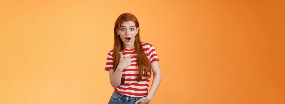 Outgoing good-looking redhead modern girl wonder making amazing purchase, look camera surprised, questioned pointing left, interested about new cool promo, stand orange background photo