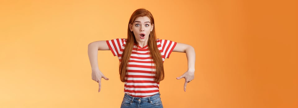 Shocked excited attractive redhead woman gasping, drop jaw shook, stare camera astonished, pointing down, feel amazed and surprised see incredible good offer, gossiping stunned, orange background photo