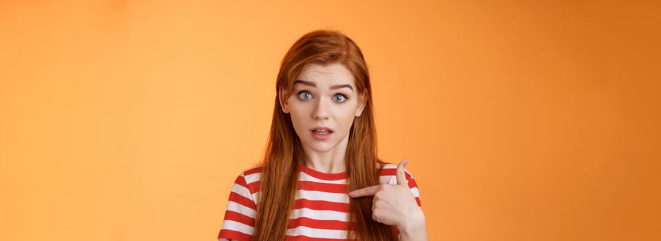 You mean me. Surprised redhead woman pointing herself puzzled raise eyebrows questioned, look full disbelief uncertain, unexpectedly hear someone mention her name, stand orange background photo