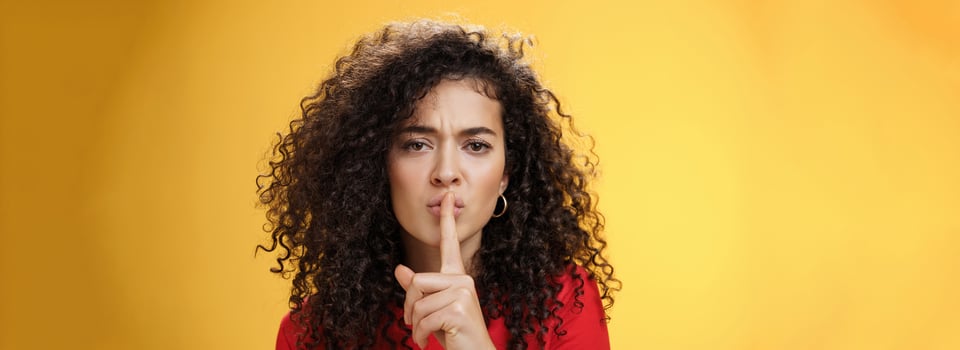 Shh not say word. Portrait of serious-looking sexy young woman with curly hair showing shush gesture with index finger over folded lips frowning asking keep secret promise keep silent over yellow wall photo
