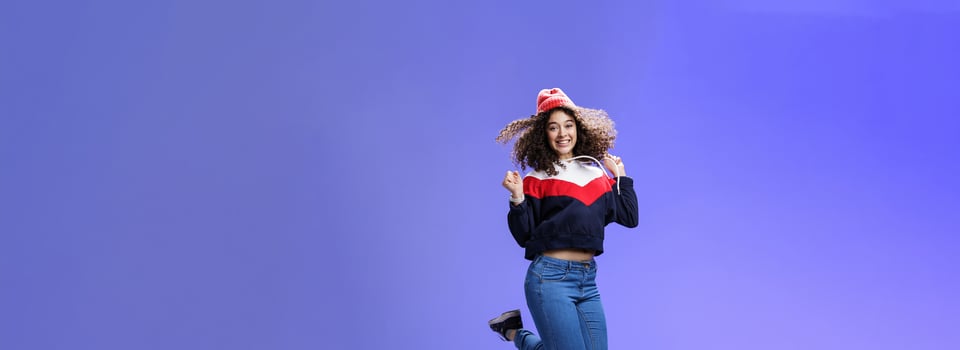 Delighted carefree and happy young woman with curly hair in warm beanie jumping joyfully having fun raising hands and smiling broadly posing over blue background in outdoor clothes photo