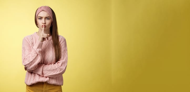 Keep mouth shut. Serious-looking bossy attractive young 20s woman in sweater, headband shushing making shhh gesture holding index finger on lips, gossiping, spread rumors over yellow background photo