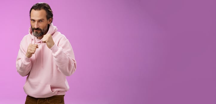Sassy confident good-looking mature caucasian bearded man in pink hoodie teaching son how stand back fight defend himself raise fists ready boxing smiling daring, stay good shape, purple background photo