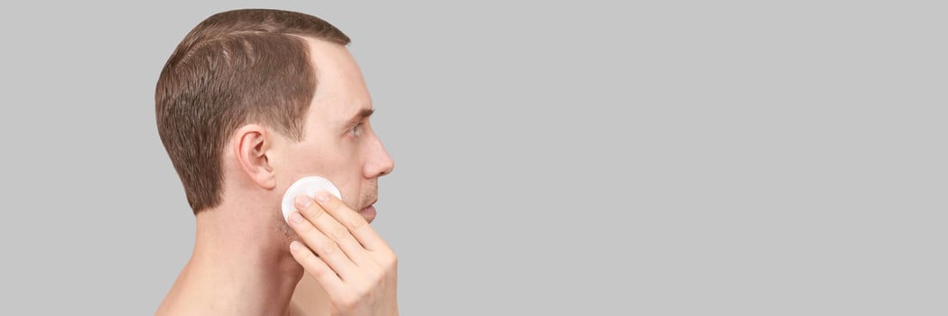 Man doing home skin care routine. Cotton pad near male chin. Dermatology treatment. Cream facial apply. Cosmetology guy portrait. Cleaning lotion. Skincare hygiene. Copyspace. Isolated on grey photo