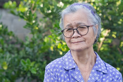 Portrait of an elderly Asian woman with short gray hair, wearing glasses, and looking away with a smile while standing outdoor. Aged people and relaxation concept photo