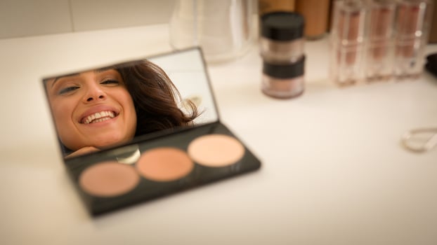 Smiling happy Caucasian woman looking reflection mirror blush palette table female girl model artist putting cosmetics makeup brush apply powder shadows visage product on face blog beauty home studio photo
